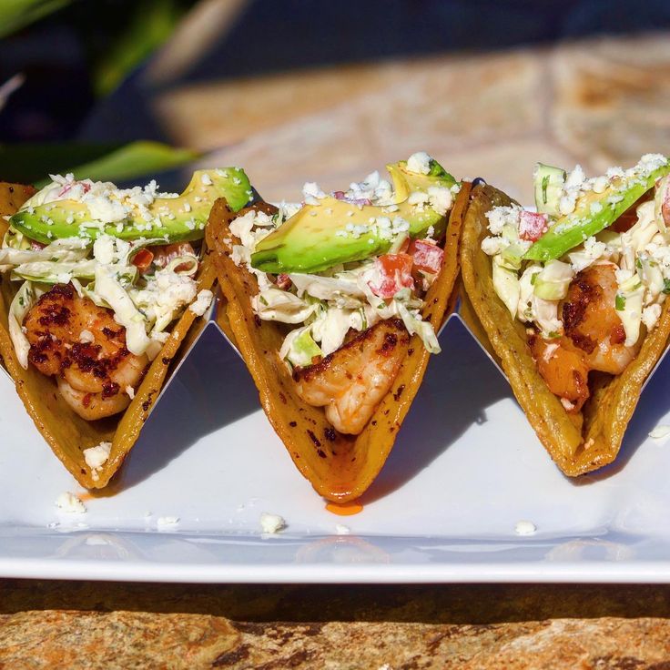 three tacos with shrimp, avocado and shredded cheese on a white plate