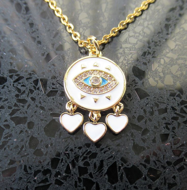 "\"Guardian of Style: Gold & White Evil Eye Pendant Necklace\" Invite a touch of mystique and protection into your jewelry collection with our Gold and White Enamel Evil Eye Pendant Necklace. 👁️ Meaningful Symbolism: The Evil Eye has been a symbol of protection across cultures for centuries. This enchanting pendant features a mesmerizing white enamel eye, bordered by radiant gold, believed to ward off negative energy and bring good fortune. ✨ Elegance Meets Protection: Crafted with precision, this necklace seamlessly blends style and symbolism. The eye's serene gaze is enhanced by the lustrous gold backdrop, creating a captivating accessory that complements any ensemble. 🛍️ Gift of Positivity: Looking for a meaningful gift? The Evil Eye necklace is not just a piece of jewelry; it's a tho Symbolic Dangle Charm Necklaces As Gift, Spiritual Charm Necklaces With Adjustable Chain, Spiritual Charm Necklace With Adjustable Chain, White Necklace With Charms And Round Pendant, White Heart-shaped Hypoallergenic Necklace, Hypoallergenic White Heart Necklace, Symbolic Charm Necklaces With Adjustable Chain, Spiritual Dangle Charm Necklaces As Gift, Spiritual Nickel-free White Jewelry