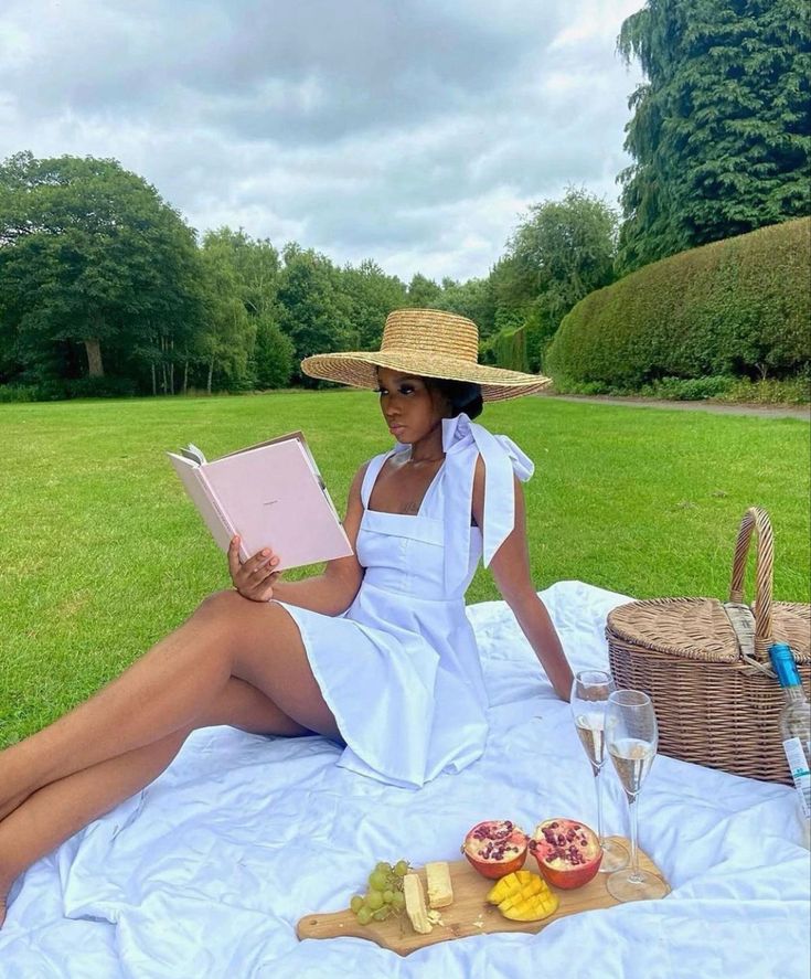 a woman in a white dress and straw hat sitting on a blanket reading a book