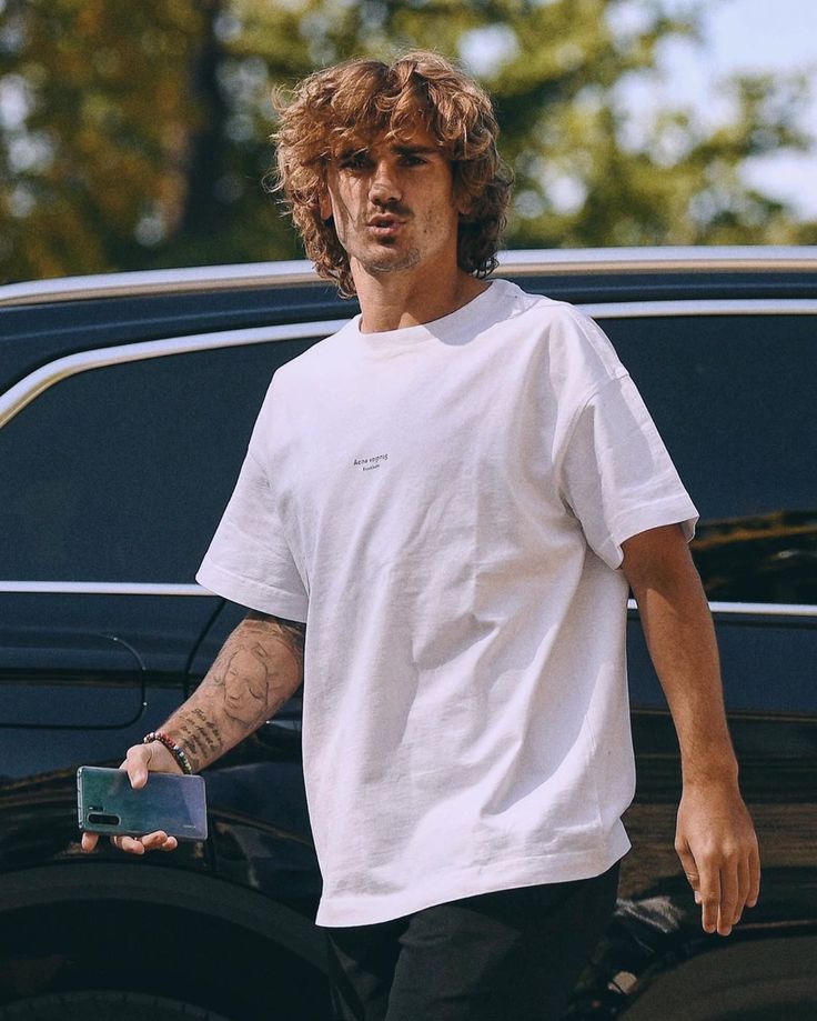 a man in white shirt and black pants walking next to a car with his cell phone