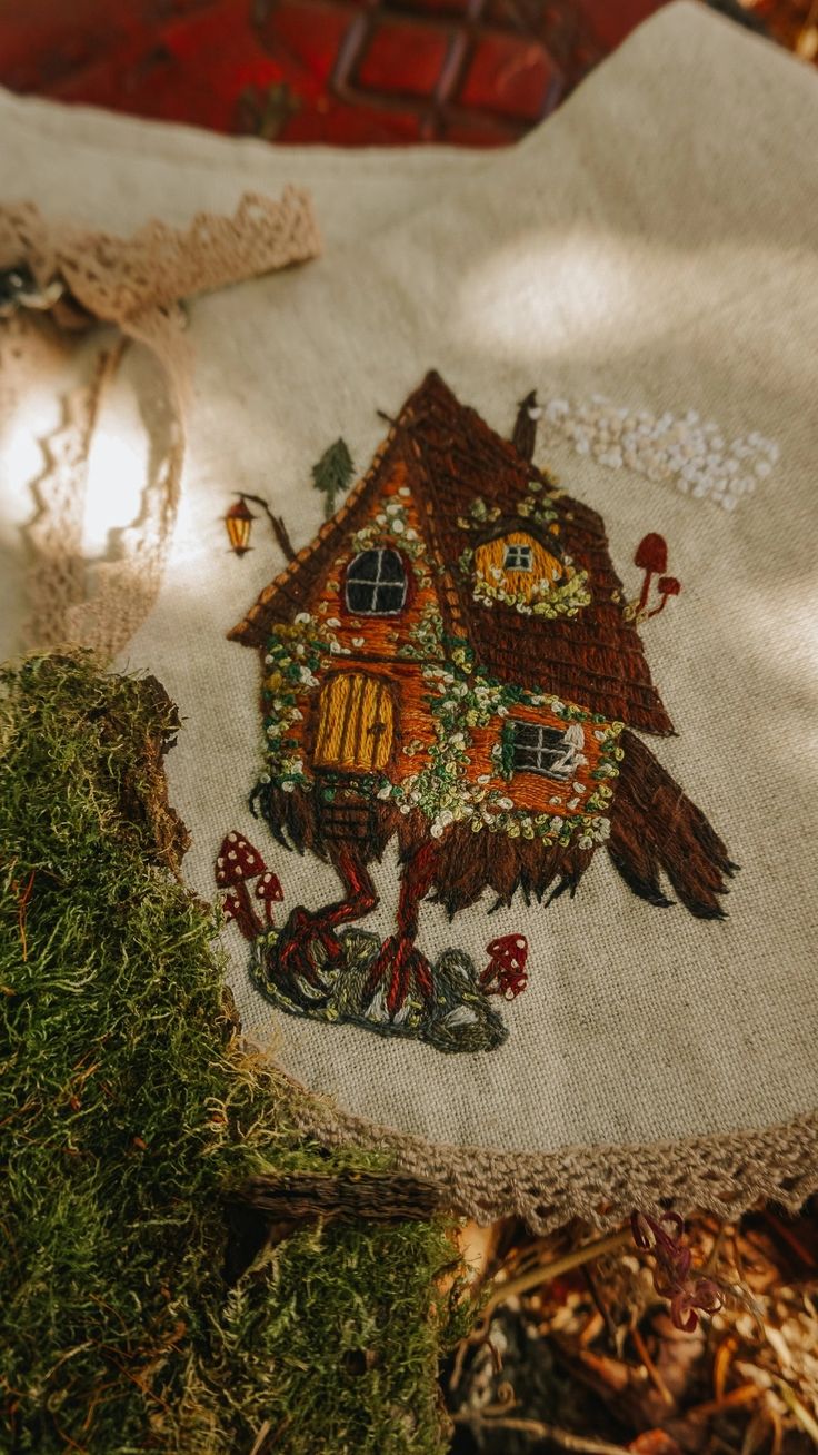 an embroidered piece of cloth with a house on it and some moss in the foreground