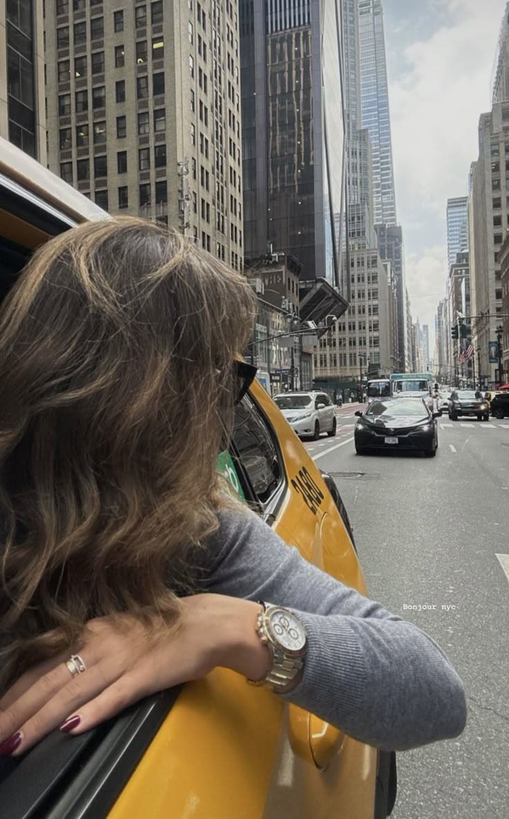 a woman leaning out the window of a taxi cab on a city street with skyscrapers in the background