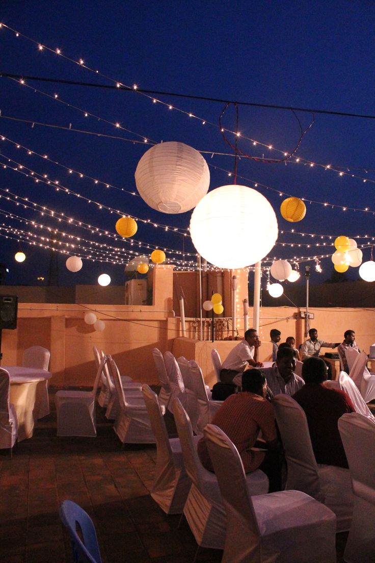 several people sitting at tables with white linens and paper lanterns hanging from the ceiling