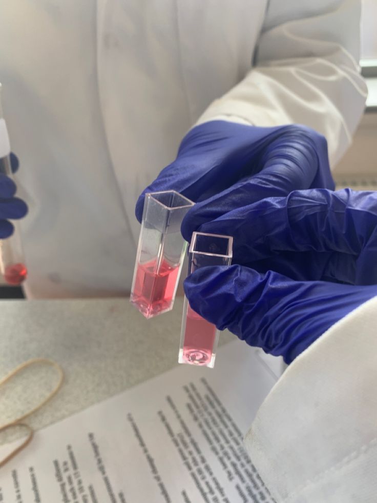 two laboratory workers in blue gloves hold test tubes filled with red liquid while looking at papers