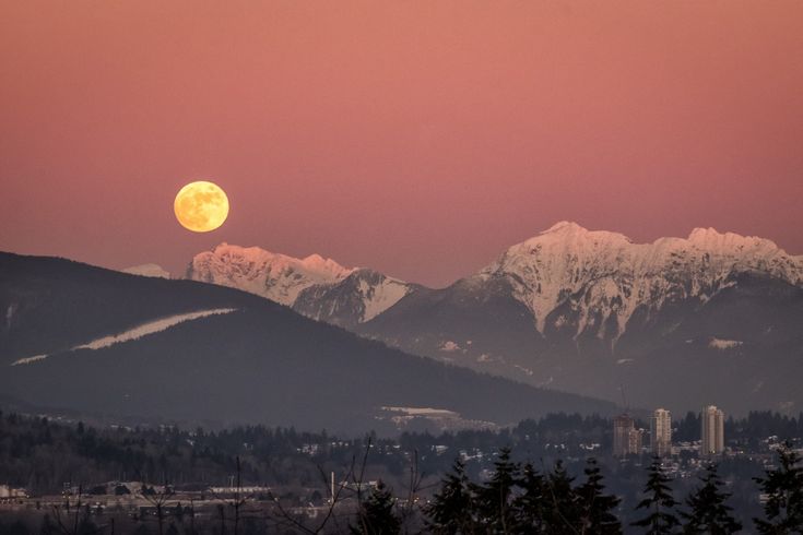 the full moon is setting over mountains with trees in foreground and snow - capped peaks