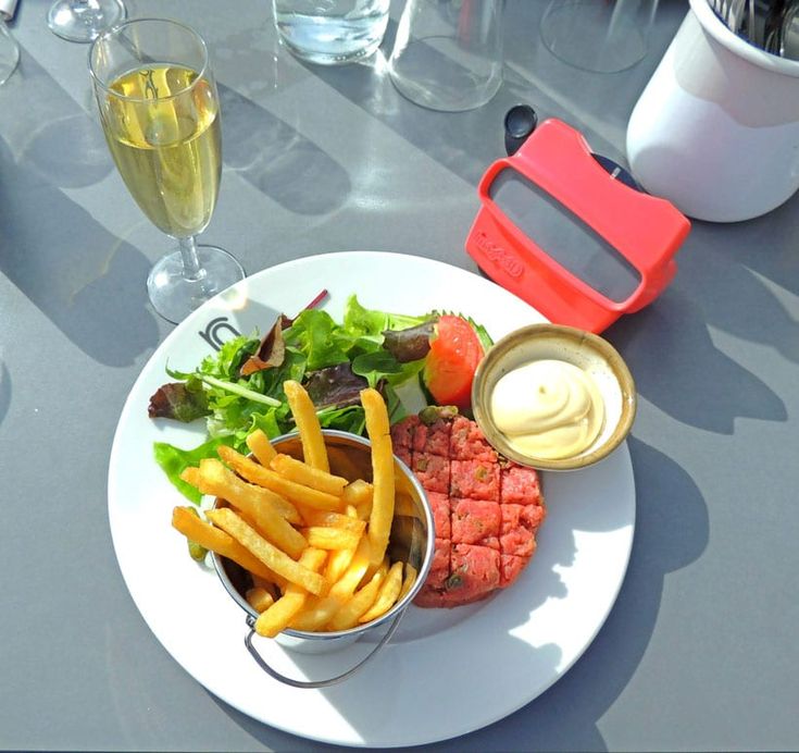 a white plate topped with meat and french fries next to a glass of wine on top of a table