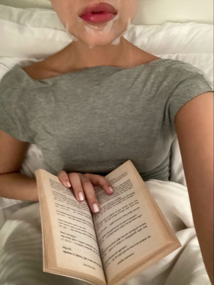 a woman laying in bed reading a book with her tongue sticking out while making a face