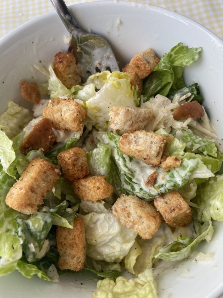 a salad with croutons and lettuce in a white bowl on a checkered table cloth