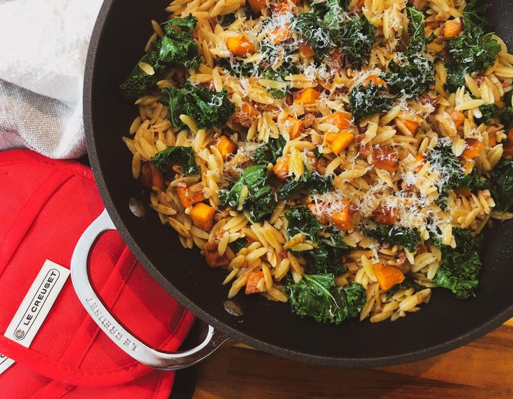 a skillet filled with pasta and vegetables on top of a wooden table next to a red towel