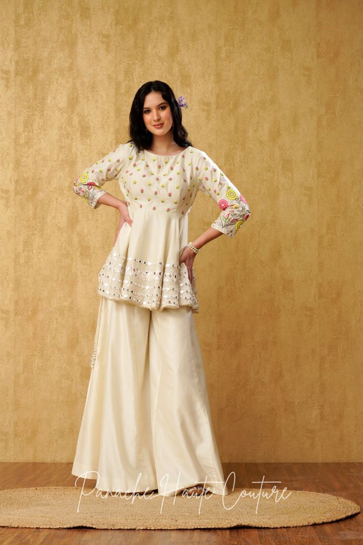 "Introducing our stunning Ivory Peplum Top with Sharara Pant, adorned with mesmerizing mirror and resham embroidery, crafted from luxurious Chanderi silk fabric." The Peplum Top is exquisitely designed to flatter the silhouette, with a chic and contemporary look. The intricate mirror and resham embroidery on the top adds a touch of elegance, creating a stunning visual effect. The top features a round neckline, complemented with a sleeveless design, which makes it perfect for any special occasion Peplum With Skirt Indian, Peplone Design, Sangeet Dress Ideas For Sisters, Cream Georgette Sharara For Reception, Festive Off-white Georgette Sharara, Beige Palazzo Set With Resham Embroidery For Reception, Cream Sets With Resham Embroidery For Reception, Cream Sharara With Resham Embroidery In Georgette, Designer Cream Sharara With Intricate Embroidery