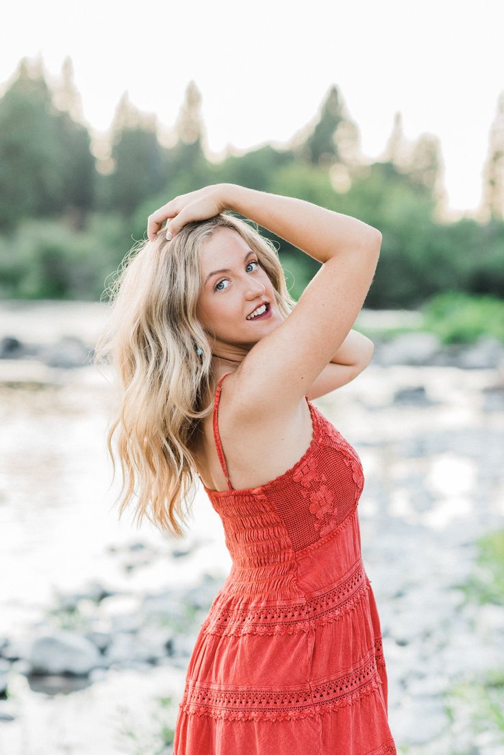 a woman in a red dress posing for a photo by the river with her hands on her head