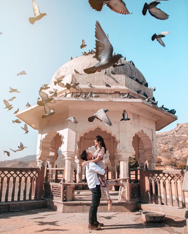 a man and woman standing in front of a building with birds flying around the structure