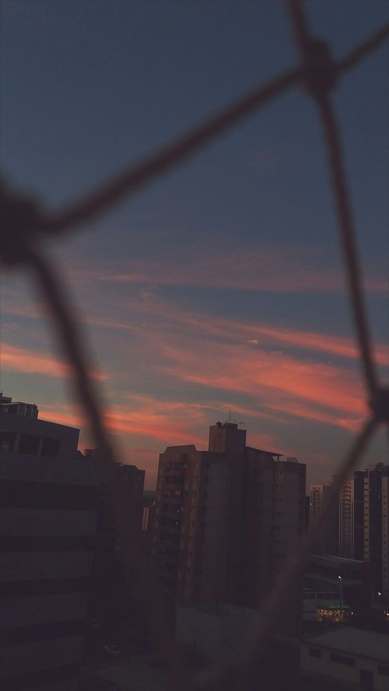 the sun is setting over some buildings and skyscrapers behind a chain link wire fence