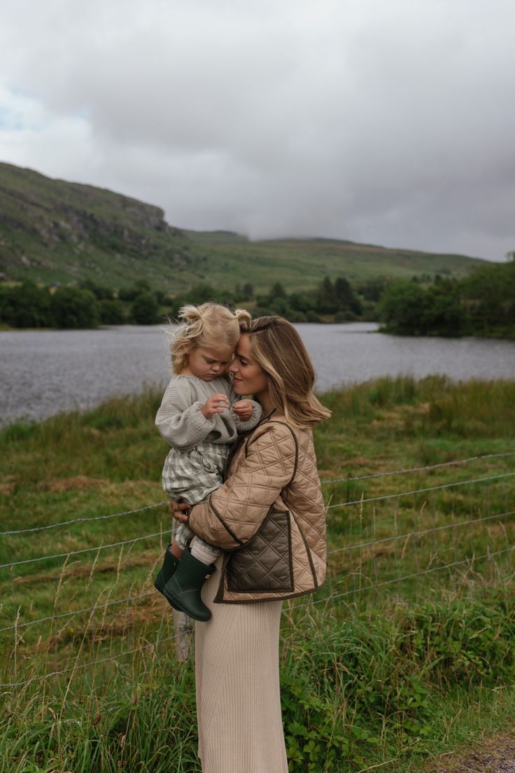 a woman holding a small child in her arms while standing next to a fence near a body of water