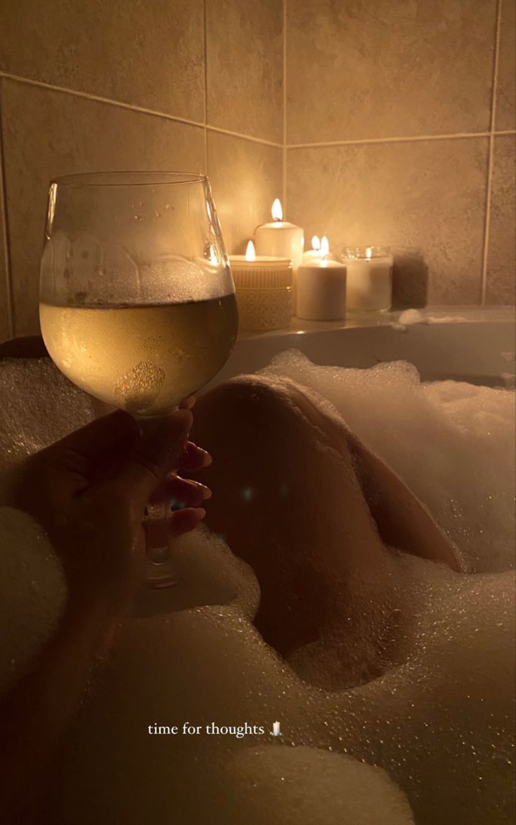 a person is taking a bath with candles in the background and wine glasses on the side