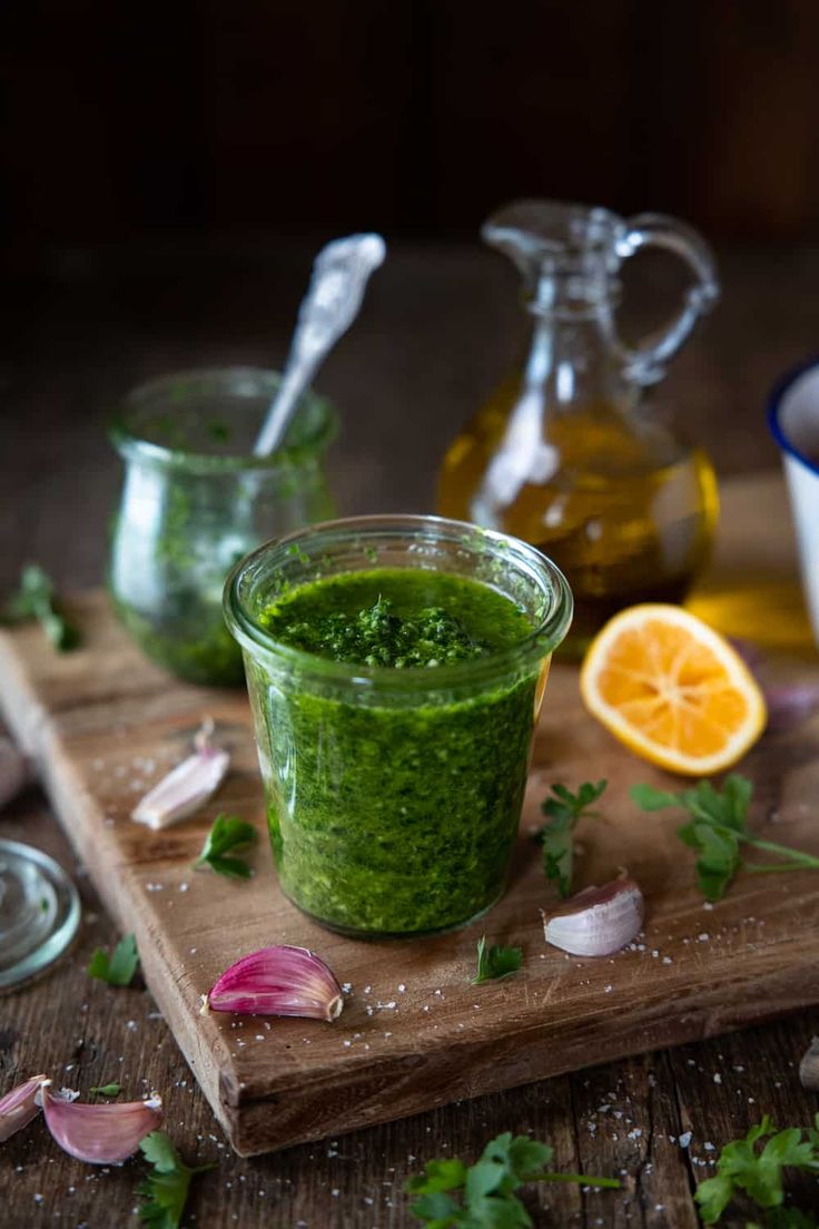 a wooden cutting board topped with a glass filled with green liquid next to garlic and an orange slice