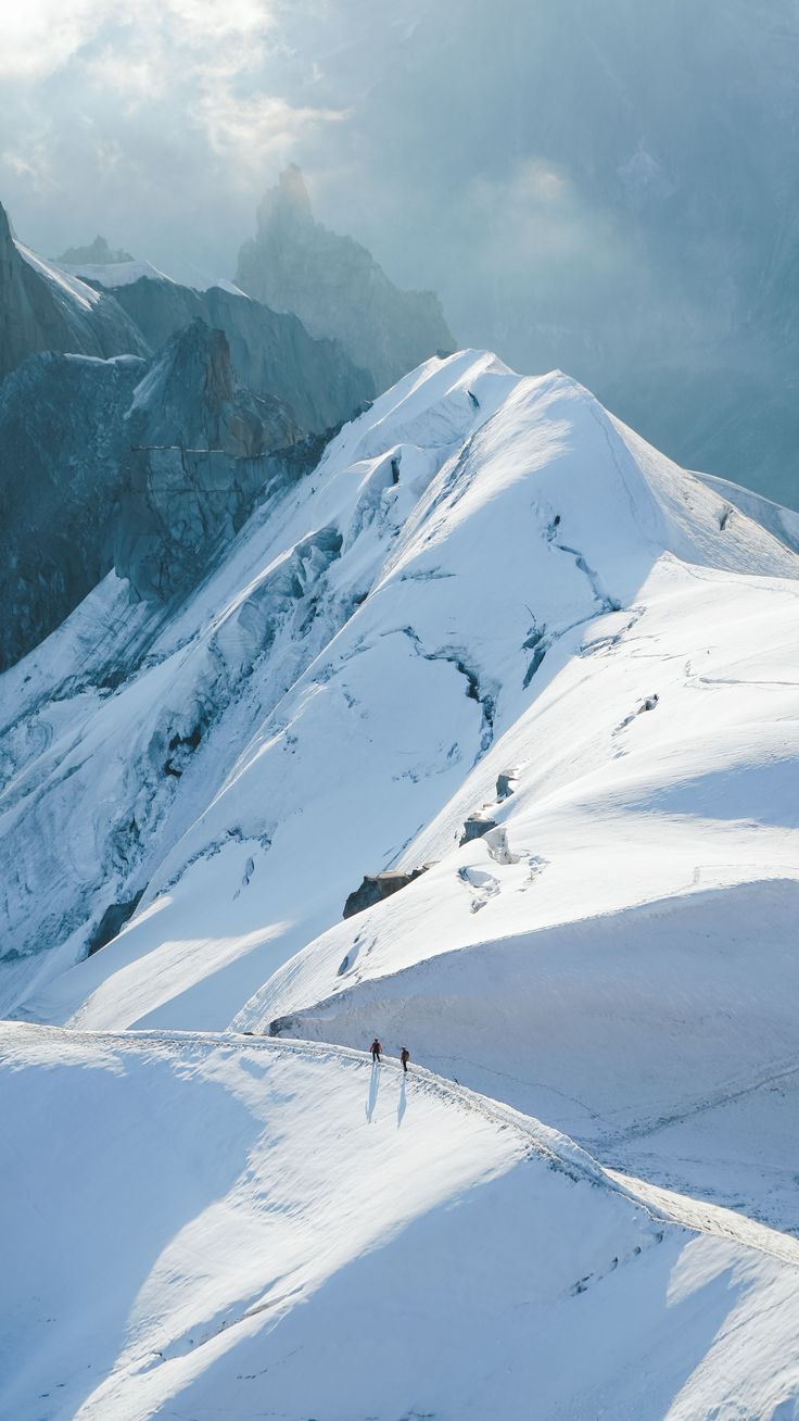 two people walking up the side of a snow covered mountain in front of some mountains