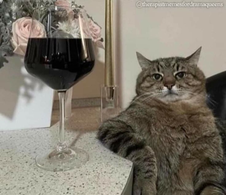 a cat sitting next to a wine glass on a table with flowers in the background