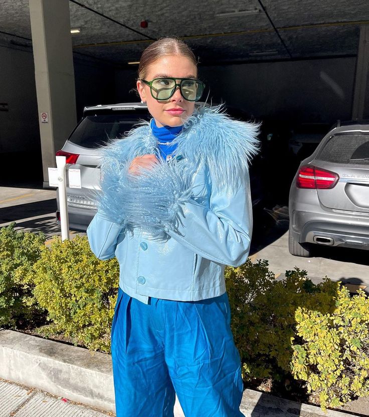 a woman standing in front of a parking lot wearing sunglasses and a blue jacket with fur on it