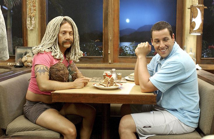 two men sitting at a table with plates of food in front of them, one man has dreadlocks on his head