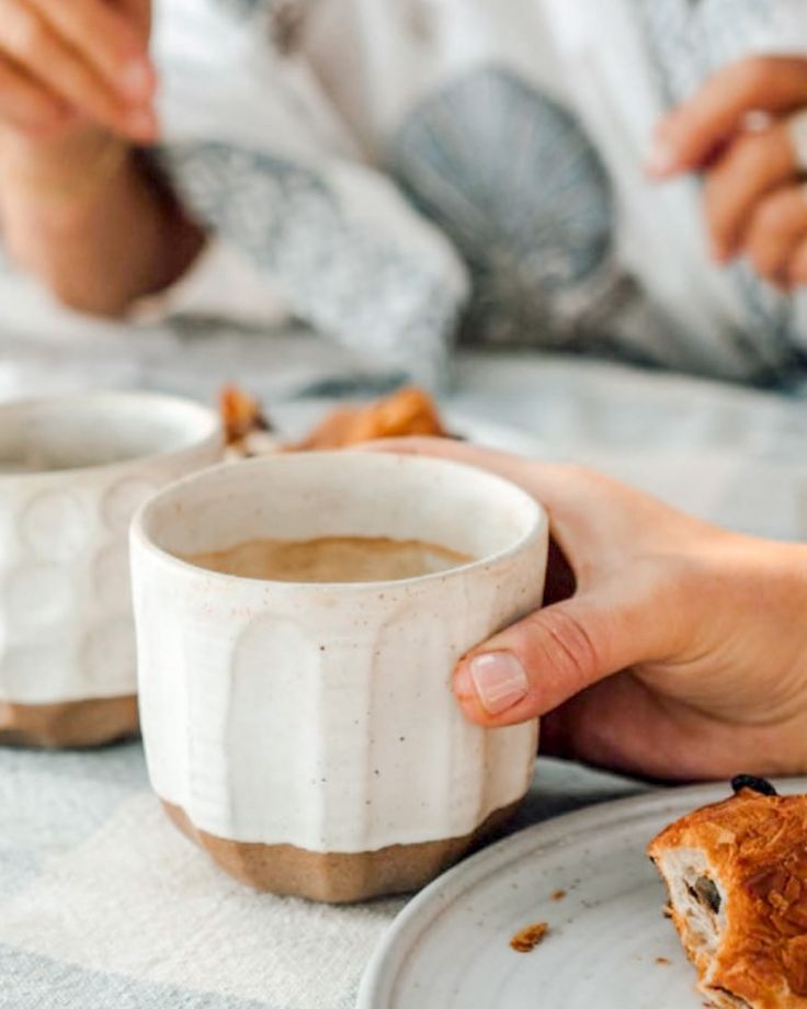 a person holding a coffee cup in their hand while sitting at a table with food