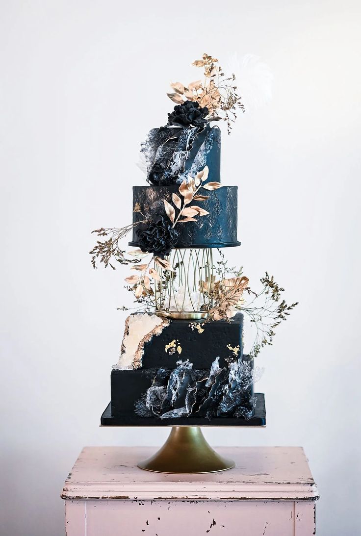 a three tiered black and gold wedding cake with flowers on the top, in front of a white wall