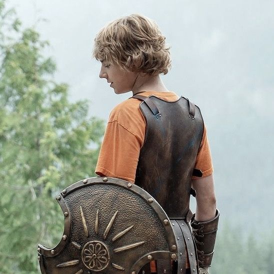 a young boy is holding a shield and looking down at the ground with trees in the background