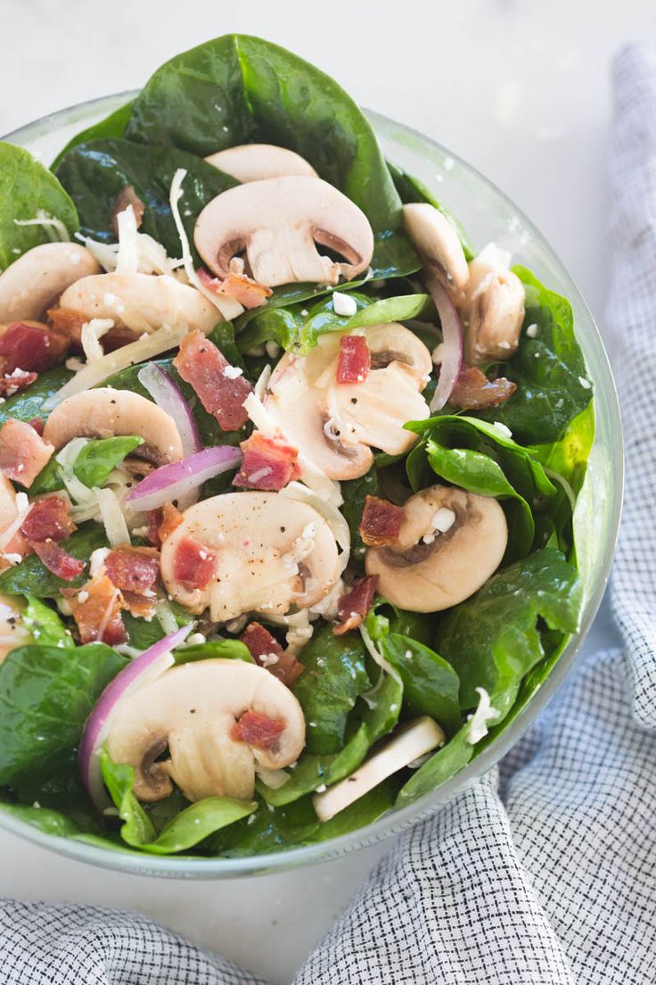 spinach salad with mushrooms and bacon in a glass bowl on top of a blue towel