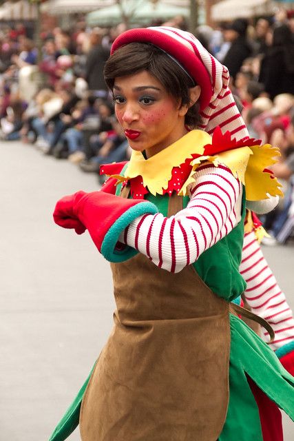 a woman dressed in costume walking down the street with people sitting on the sidelines behind her