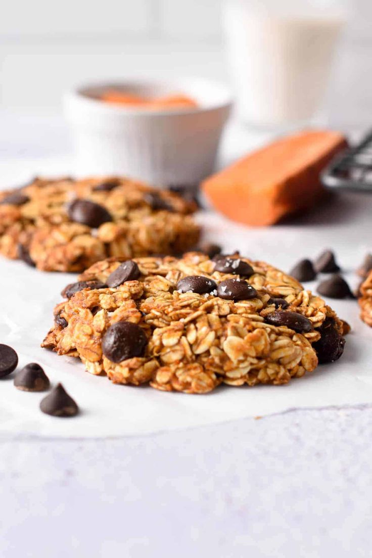 three cookies with chocolate chips and oats on a white surface next to a glass of milk