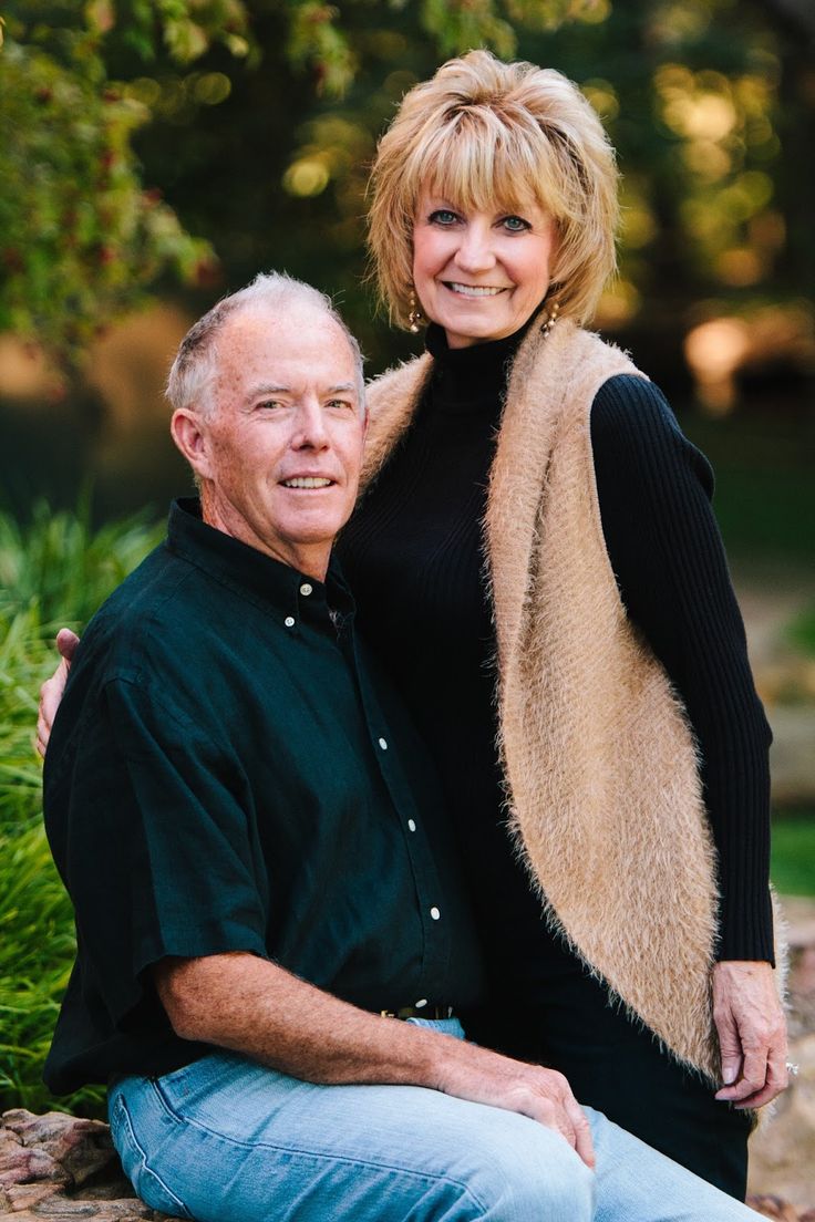 an older man and woman sitting next to each other in front of some bushes, smiling at the camera