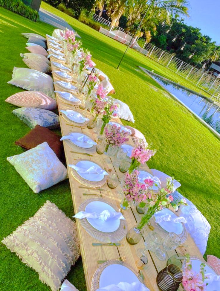 a long table set up with plates and place settings for dinner on the grass near water