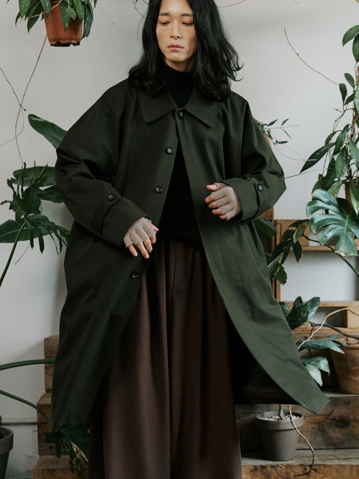 a woman standing in front of some plants wearing a green coat and brown wide legged pants