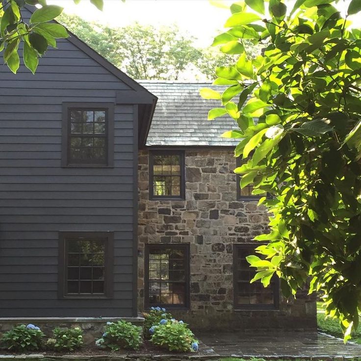 a stone house with black shingles on the front and side walls, surrounded by greenery