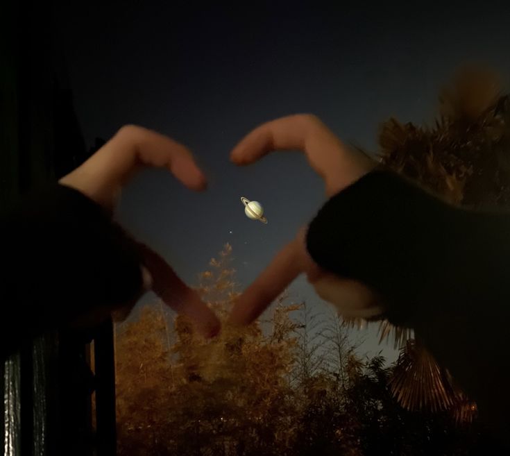 two hands making a heart shape with the moon in the sky and trees behind them