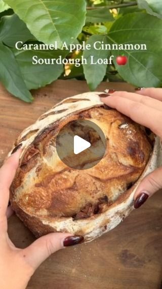 a person holding a loaf of bread on top of a wooden table next to green leaves