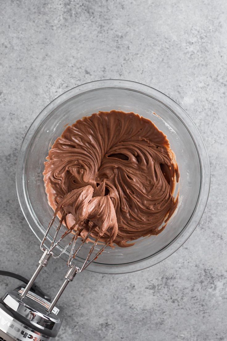 chocolate frosting in a glass bowl with a whisk on the side and an electric hand mixer next to it