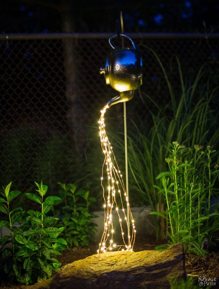 a metal watering can with fairy lights on it in the middle of a garden at night