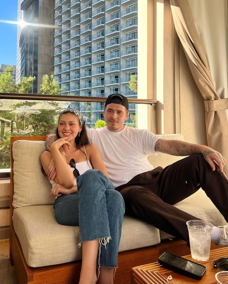 a man and woman sitting on a couch in front of a window with the city behind them