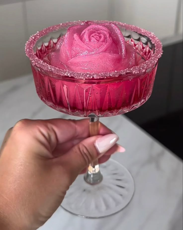 a hand holding a wine glass with pink liquid in it and a rose on the rim