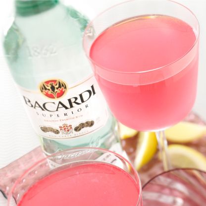 a glass filled with pink liquid next to a bottle of bacardi water and lemon wedges