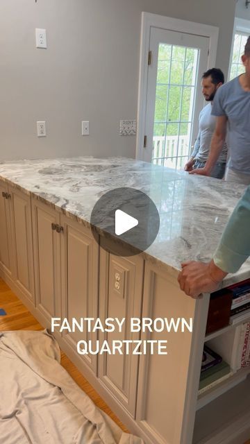 two men standing in a kitchen next to an island with marble counter tops and drawers