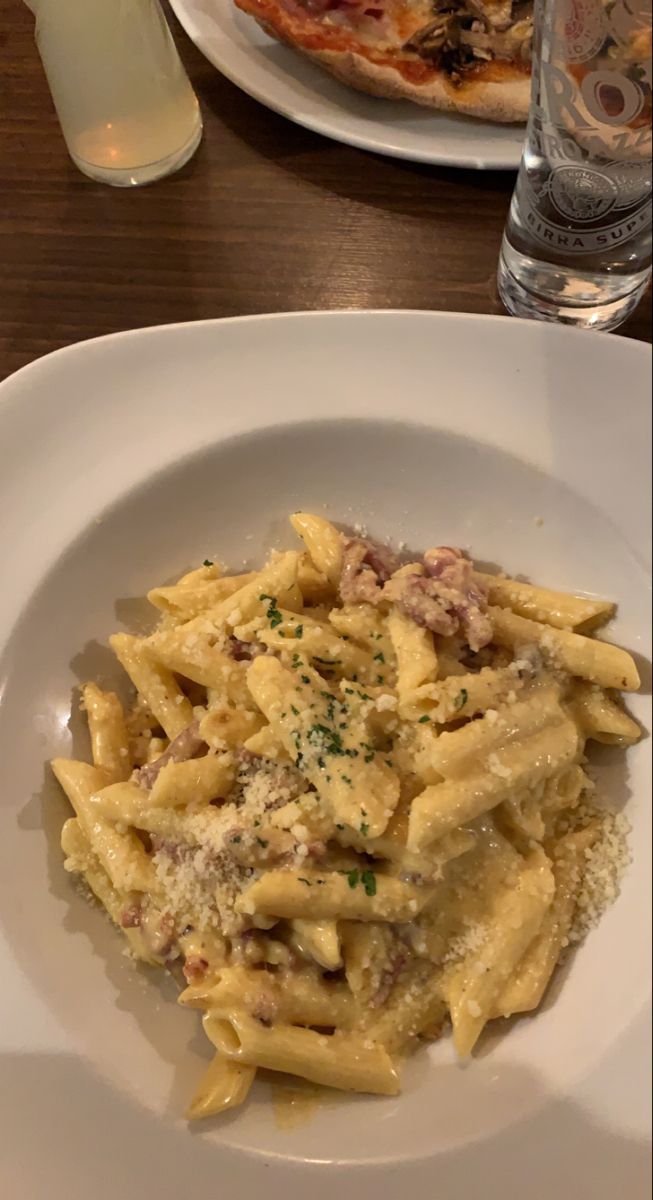 a white plate topped with pasta and meat covered in sauce next to a slice of pizza