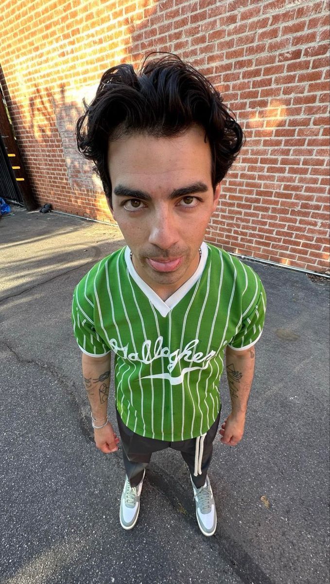 a man with black hair wearing a green and white baseball jersey standing in front of a brick wall