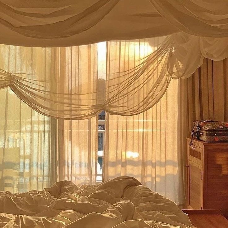 an unmade bed sitting in front of a window with sheer drapes on it