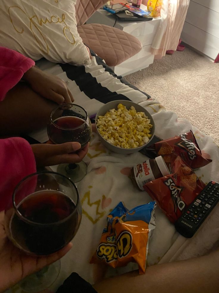 two people sitting on a bed with snacks and drinks in front of them, one holding a glass of wine