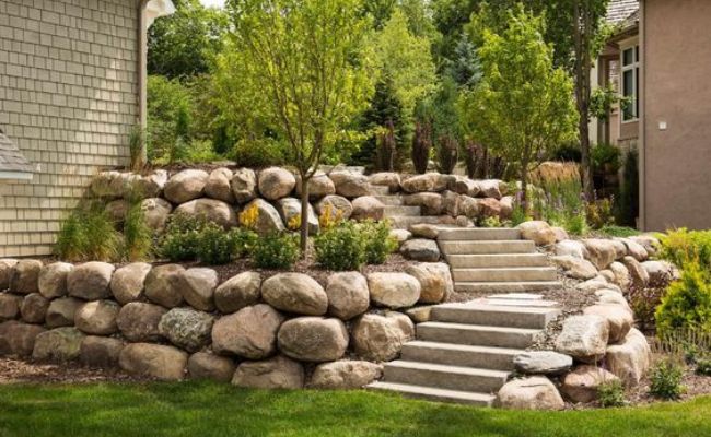 a stone wall next to a house with steps leading up to the front door and landscaping