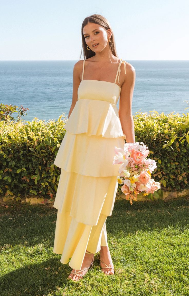 a woman standing in the grass wearing a yellow tiered dress and holding a bouquet