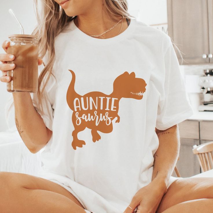 a woman sitting on a bed holding a drink and wearing an orange dinosaur shirt with the words,'auntiesauruss'printed on it