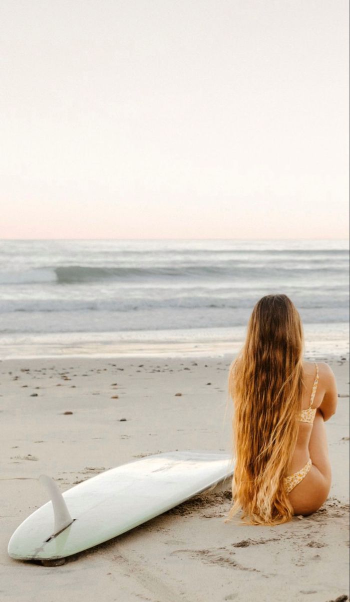 a woman sitting on the beach next to a surfboard looking out at the ocean